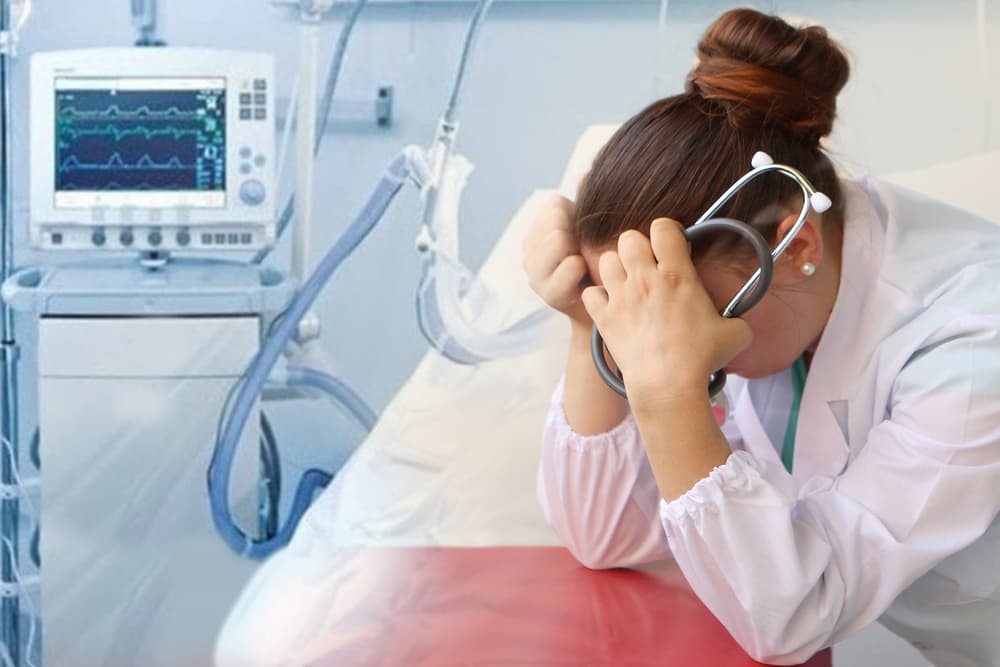 Female doctor with a stethoscope covering her head with her hands, next to a medical ventilator, illustrating despair and medical fatigue.