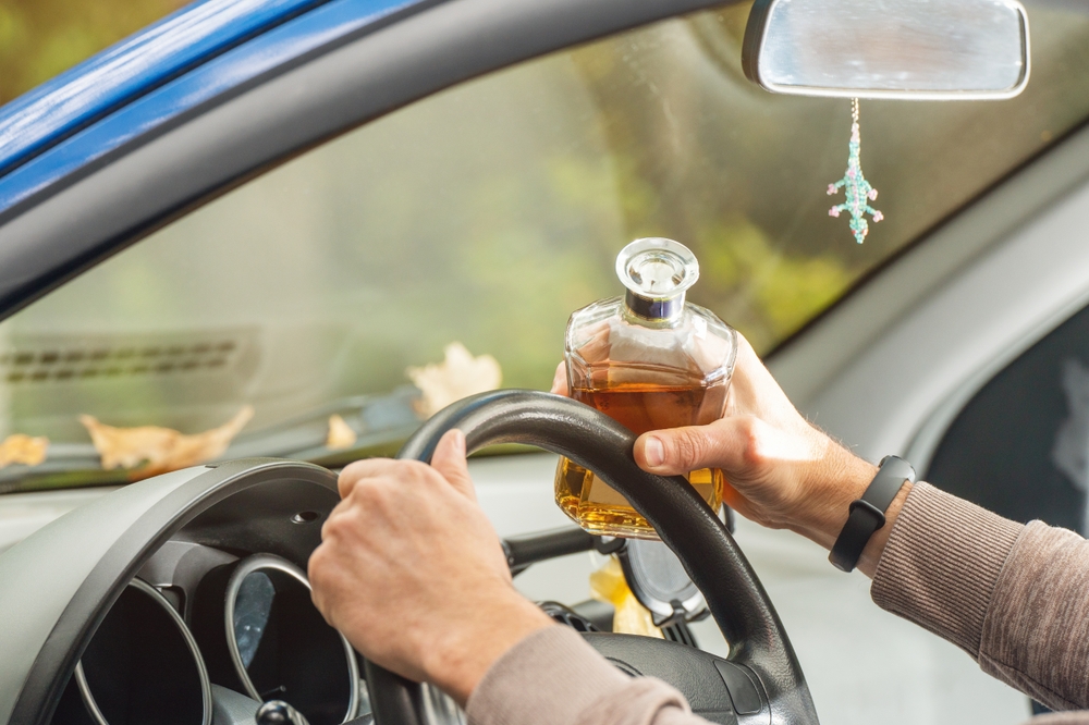 The driver is behind the wheel, holding a bottle of alcohol, breaking the law by drinking while driving.