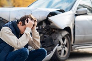 A frustrated man stands by a broken car, holding his head as he realizes the damage is severe and the car is beyond repair.