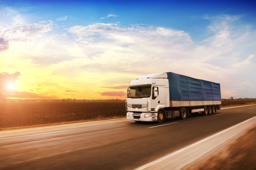 Large white truck with a blue trailer driving on a countryside road at sunset, with a blank space for text against an evening sky.