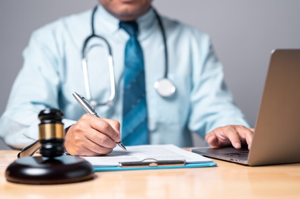 A doctor with a stethoscope writes on documents while using a laptop, with a gavel nearby.