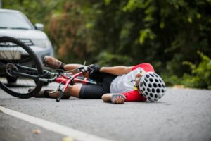 A cyclist wearing a helmet lies injured on the road after falling, possibly suffering a degloving injury.