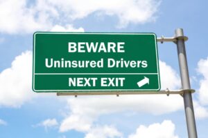 Green road sign reads "Suing Uninsured Drivers? Next Exit" against a partly cloudy sky.